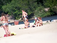 Frisky man with the working camera is lying on the hot golden sand of the nudist beach voyeuring sexy nude body from the distance